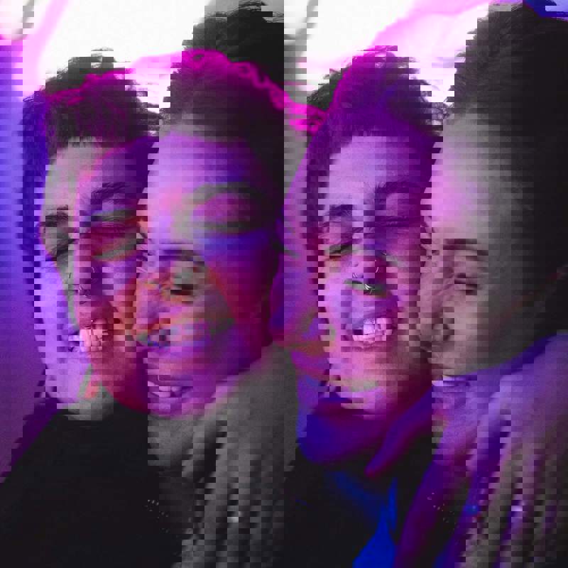 Two women smile and embrace warmly. The image is bathed in a pink and purple light.