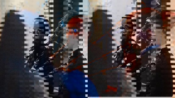 Two men are playing bagpipes indoors. One man has a beard and glasses, and the other is smiling.