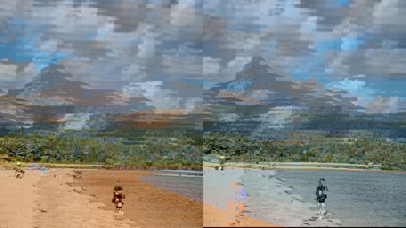 Two walkers stroll along a sandy beach bordering a calm bay, with the distinctive peak of Goat Fell and dense green forests rising in the background.