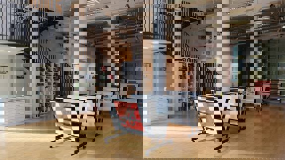 An empty art studio and gallery with a light wooden floor. The space has a staircase, white support columns, and racks displaying framed artwork.