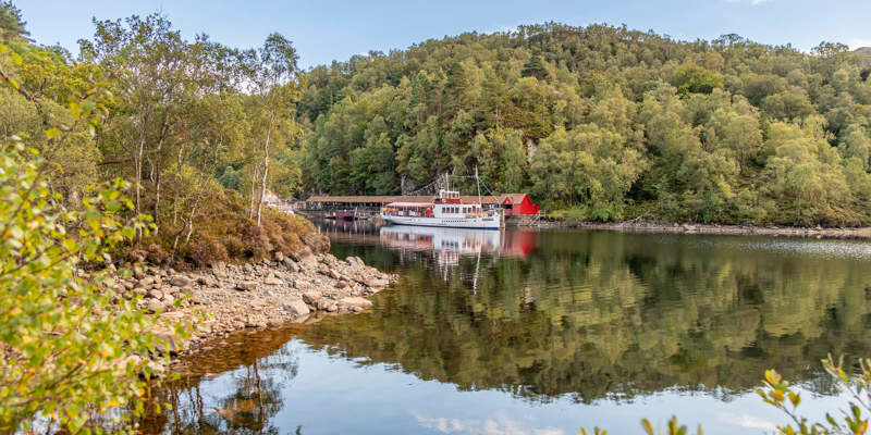 A tranquil view of Loch Katrine's Trossachs Pier, featuring a classic white steamship reflected in the calm water.