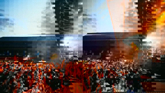 A lively crowd with raised hands watches a musician perform on an outdoor stage at SWG3 in Glasgow, bathed in warm orange light.