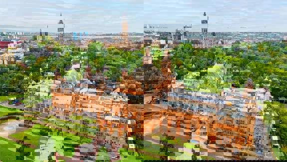 Drone view of a grand red building surrounded in greenspace, with a grand building with a spire and hills in the distance.