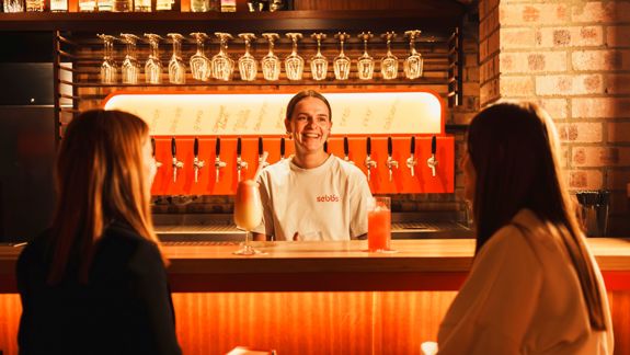 A smiling bartender stands behind a warmly lit bar with exposed brick walls and a row of beer taps, serving two women seated at the counter. Colourful cocktails sit on the bar, with hanging glassware and bottles displayed on shelves above, creating a cosy, inviting atmosphere.