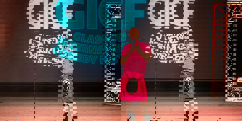 A smiling comedian in a pink shirt and kilt is on stage at the Glasgow International Comedy Festival (GICF) with a microphone.