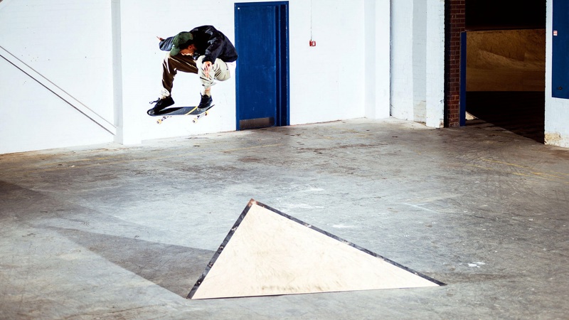  Skateboarder performing an ollie off a triangular wooden ramp in an empty, industrial indoor space. 