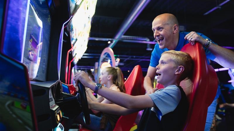 An adult and child are enthusiastically playing a racing arcade game, both looking excited and focused on the screen.
