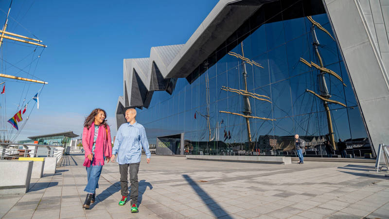 Two people walk and talk outside a modern museum with a striking zigzag roof and glass walls reflecting a tall ship under a clear blue sky.