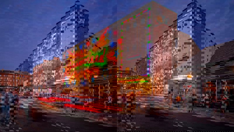 A street-level view of the Barrowland Ballroom at night. The building is lit with a colourful neon sign with stars and the name "Barrowland" in a large font. The long exposure shot shows blurred light trails from cars on the street.