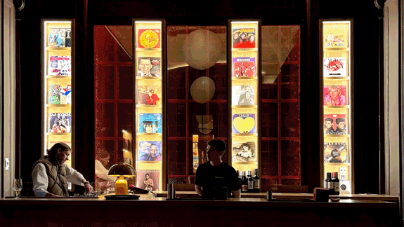 A DJ and bartender stand behind a bar with tall, backlit shelves of vinyl records behind them.