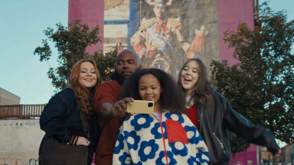A group of four smiling friends take a selfie together outdoors. They stand before a pink building featuring a large mural, surrounded by green trees under a clear sky.