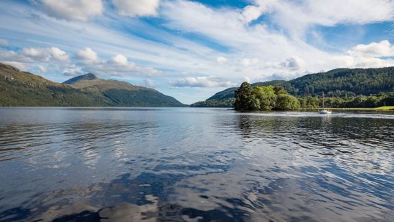 A serene view of Loch Lomond, Scotland, with calm water reflecting the blue sky and white clouds, surrounded by green hills. A small boat is visible on the right.