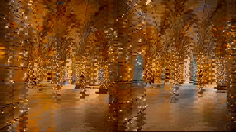 The arched stone cloister of the University of Glasgow adorned with strings of golden fairy lights in the evening.