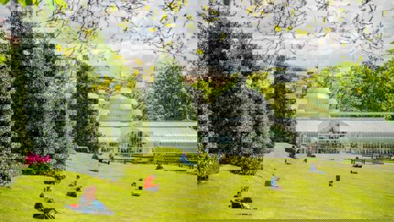 People sitting on the lawn at Glasgow Botanic Gardens, with the domed glasshouse of Kibble Palace in the background.