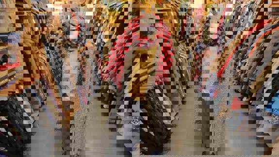 Interior view of a vintage clothing store with wooden shelving units flanking a central aisle. A mannequin in the middle wears a tweed coat, a yellow shirt, and a red and black checkered scarf.