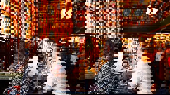 A couple reviews a menu at a table in a bar lined with bookshelves, illuminated by warm lighting from lamps and shelves.