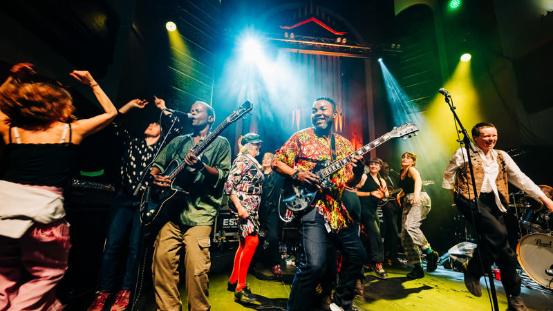 A lively band and backing dancers perform on a brightly lit stage, with one guitarist smiling and playing.