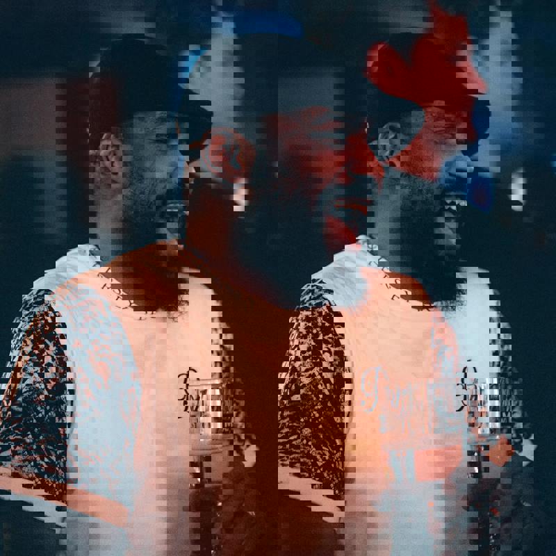 A smiling man in a black cap and white patterned shirt holds a glass of beer. He is laughing, and other people are visible in the blurred background.