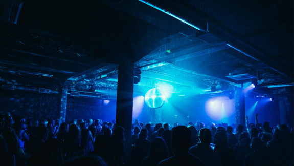 Crowd dancing in a large, dark warehouse venue with a mirror ball reflecting blue light beams across the room, creating a vibrant nightlife atmosphere.