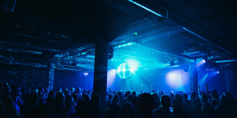 Crowd dancing in a large, dark warehouse venue with a mirror ball reflecting blue light beams across the room, creating a vibrant nightlife atmosphere.