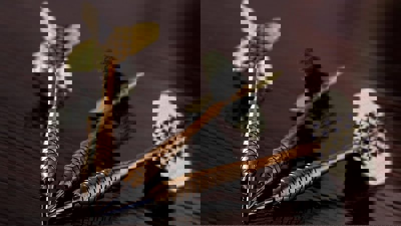 Three metallic darts with patterned flights lying on a polished wooden surface, arranged in a close cluster with tips pointing inward.