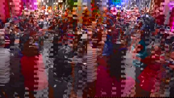 A diverse group of people of all ages holding hands in a large circle, celebrating and dancing in a brightly lit hall or function room.