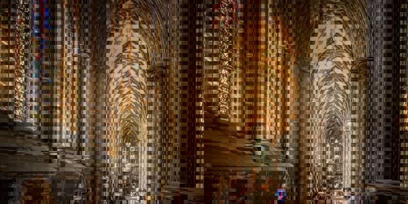 A long, vaulted cathedral nave with stone columns and stained glass windows, light streaming in from the right.