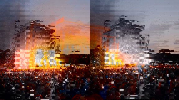 A night time crowd holding up phones to capture a performance on a large, brightly-lit stage with pyrotechnics.