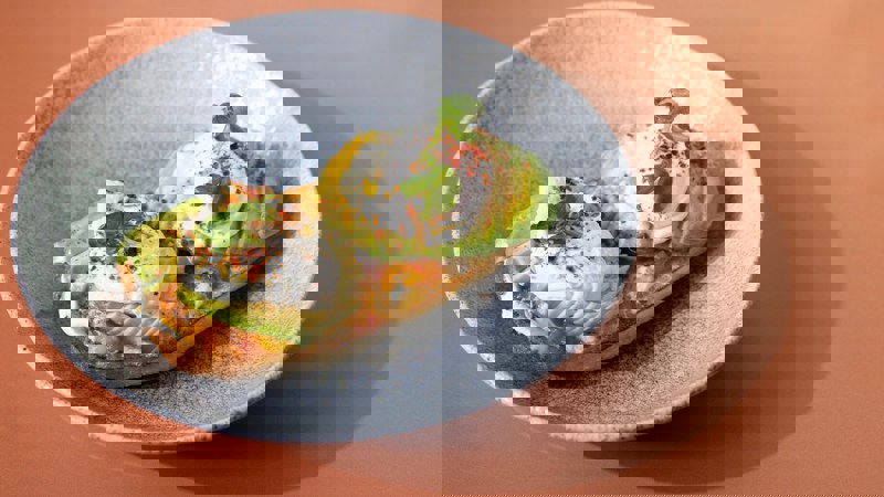 Close-up of a brunch dish of avocado toast with poached eggs and chilli flakes on a grey plate against a peach coloured table.