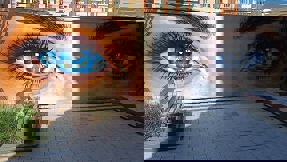 Close-up mural of large, vivid blue eyes on a building wall next to a paved area with benches.