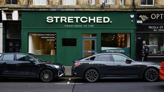 A wellness studio named "STRETCHED. FROM HEAD TO TOE" with a dark green facade is visible between two parked dark grey cars on a city street. The sign lists services like 1-ON-1 ASSISTED STRETCHING, INFRARED SAUNA, and ICE BATHS.