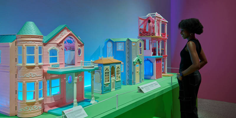 A woman views an exhibition display featuring several large, colourful Barbie Dreamhouses from different eras, illuminated by bright blue and pink lights.
