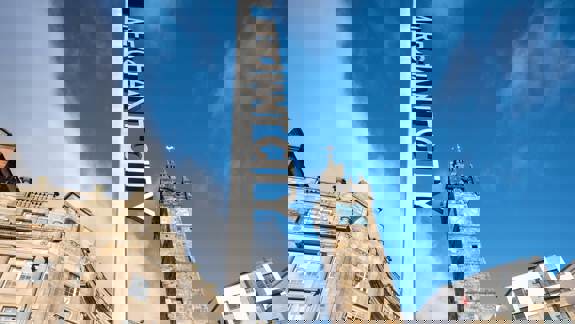 A modern, tall sign with the words 'Merchant City' on it, standing in front of the historic Tolbooth Clock Tower in Glasgow.