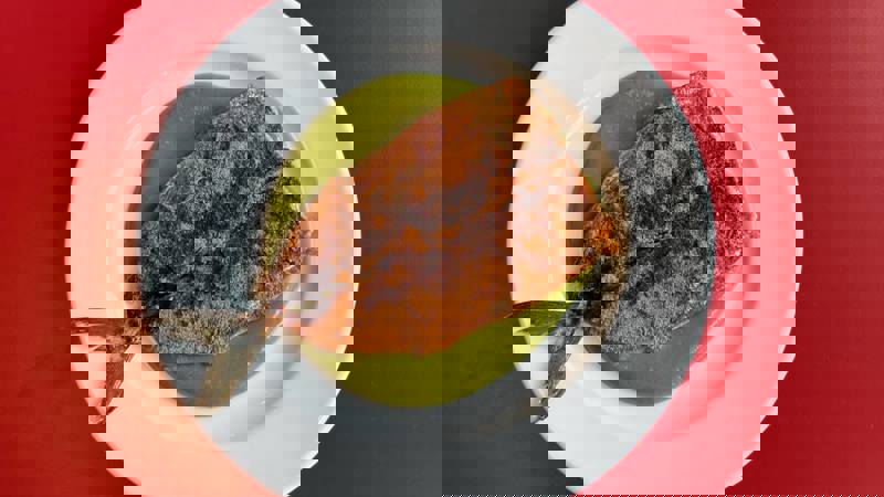 A seared mackerel fillet with a tail is served on a white plate with a green sauce. The plate is on a red table.