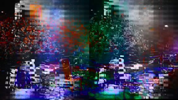 An audience seated in a dark theatre watches a performance on a smoke-filled stage lit with colourful lights, with musical instruments in the foreground.