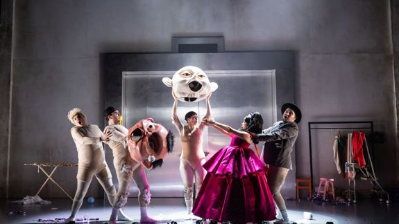 Five performers in a theatrical scene hold oversized masks on a dim stage. One wears a vibrant pink dress, creating a surreal, whimsical atmosphere.