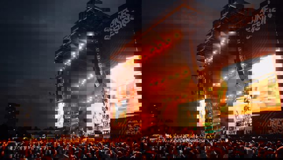 Large outdoor concert stage at Summer Sessions festival, brightly lit with screens and performers, as a crowd gathers under a dramatic evening sky