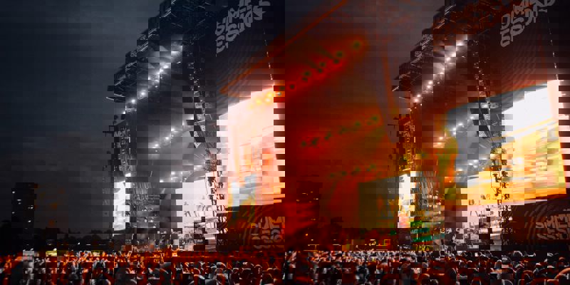 Large outdoor concert stage at Summer Sessions festival, brightly lit with screens and performers, as a crowd gathers under a dramatic evening sky