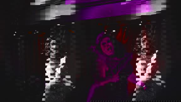 A drag queen with glitter makeup and a sparkly outfit performs on a stage, holding a fan.