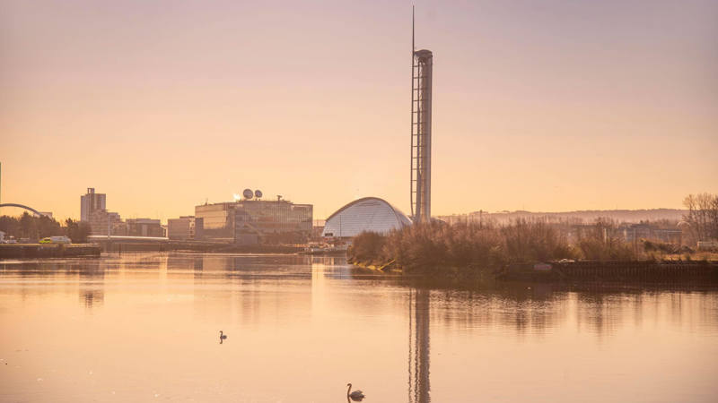 A calm river at sunset with swans swimming, modern buildings, and a tall tower in the background.