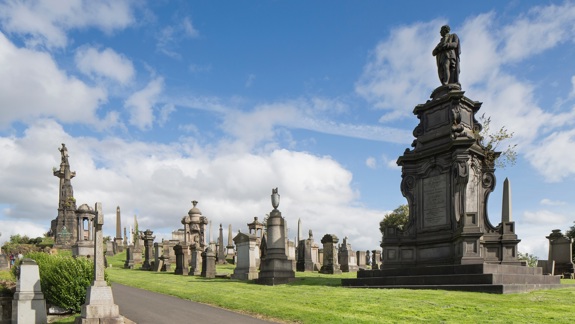 Glasgow Necropolis is filled with ornate stone monuments and statues under a bright blue sky with scattered clouds.