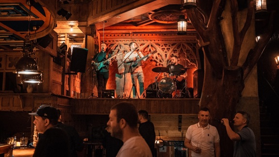 A band plays on a stage in a pub with people gathered to watch the performance. The bar has a dark, wood interior, with a prominent tree trunk feature on the right side of the image. 