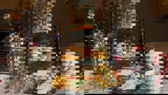 A three-tiered stand with sandwiches, chips, scones, and cake. Two glasses of fizz, a teapot, and a floral setting create a vintage afternoon tea scene.