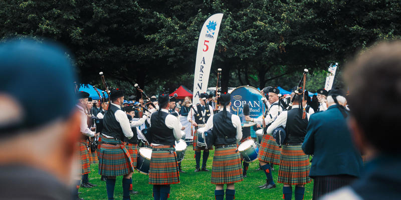 A pipe band, dressed in kilts and jackets, performs a formation on the grass, surrounded by an audience.