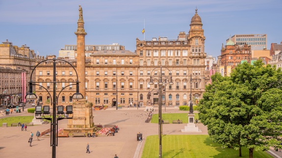 It's a sunny day in George Square where citizens are going about their day. Glasgow City Chambers and the Sir Walter Scott statue are the centre of the shot. 
