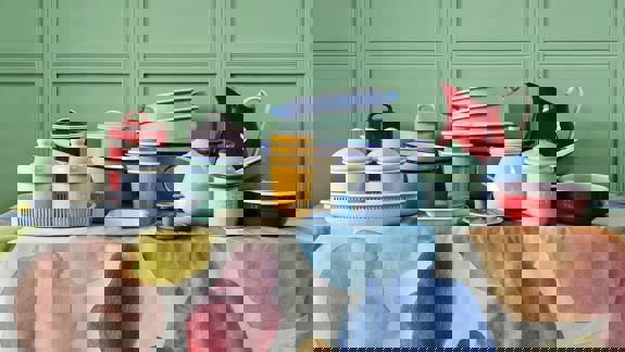 A collection of pastel and colourful enamel kitchenware, including a pitcher, cups, and bowls on a patterned tablecloth.
