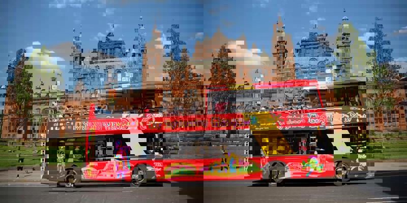 A bright red double-decker city sightseeing bus in Glasgow, with passengers on the top deck, passing the grand Kelvingrove Art Gallery and Museum.