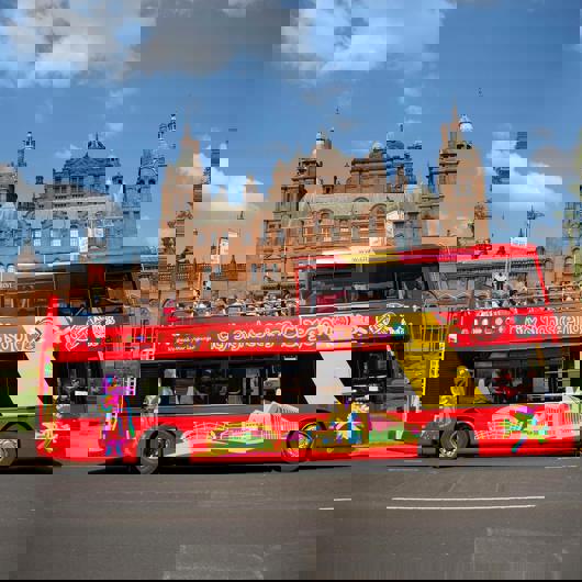 A bright red double-decker city sightseeing bus in Glasgow, with passengers on the top deck, passing the grand Kelvingrove Art Gallery and Museum.