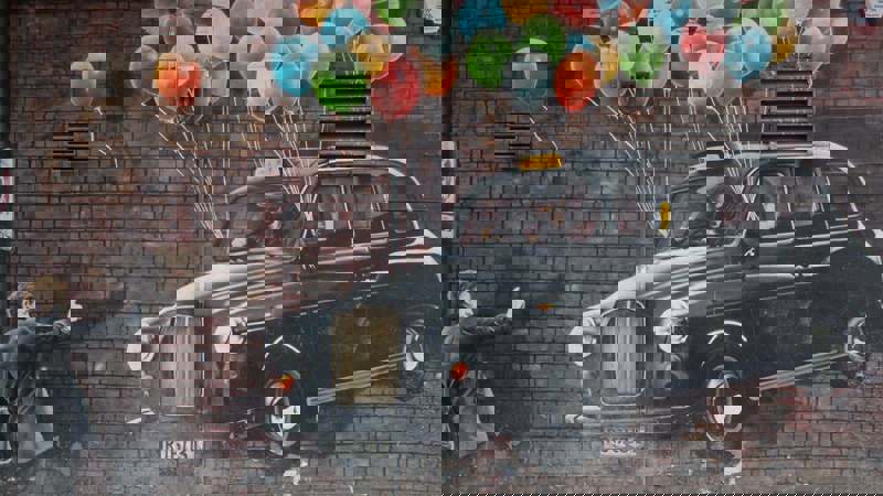 A mural on a brick wall showing a black taxi floating upwards, held by a large bunch of colourful balloons.