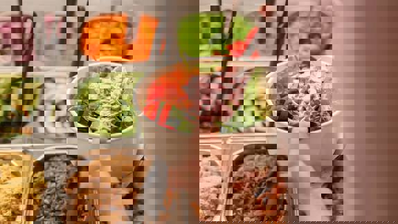 A person holds a white bowl filled with a fresh salad, including avocado and a pink sauce, over a buffet of colourful ingredients.